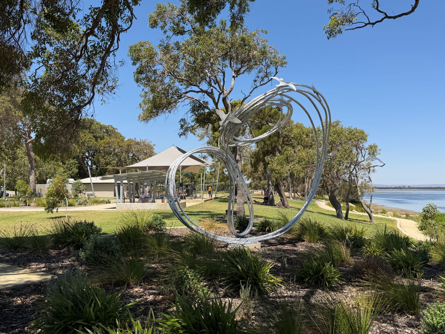 A large silver metal sculpture of swirling loops and flying birds stands in a grassy park beside an estuary on a clear, sunny day.
